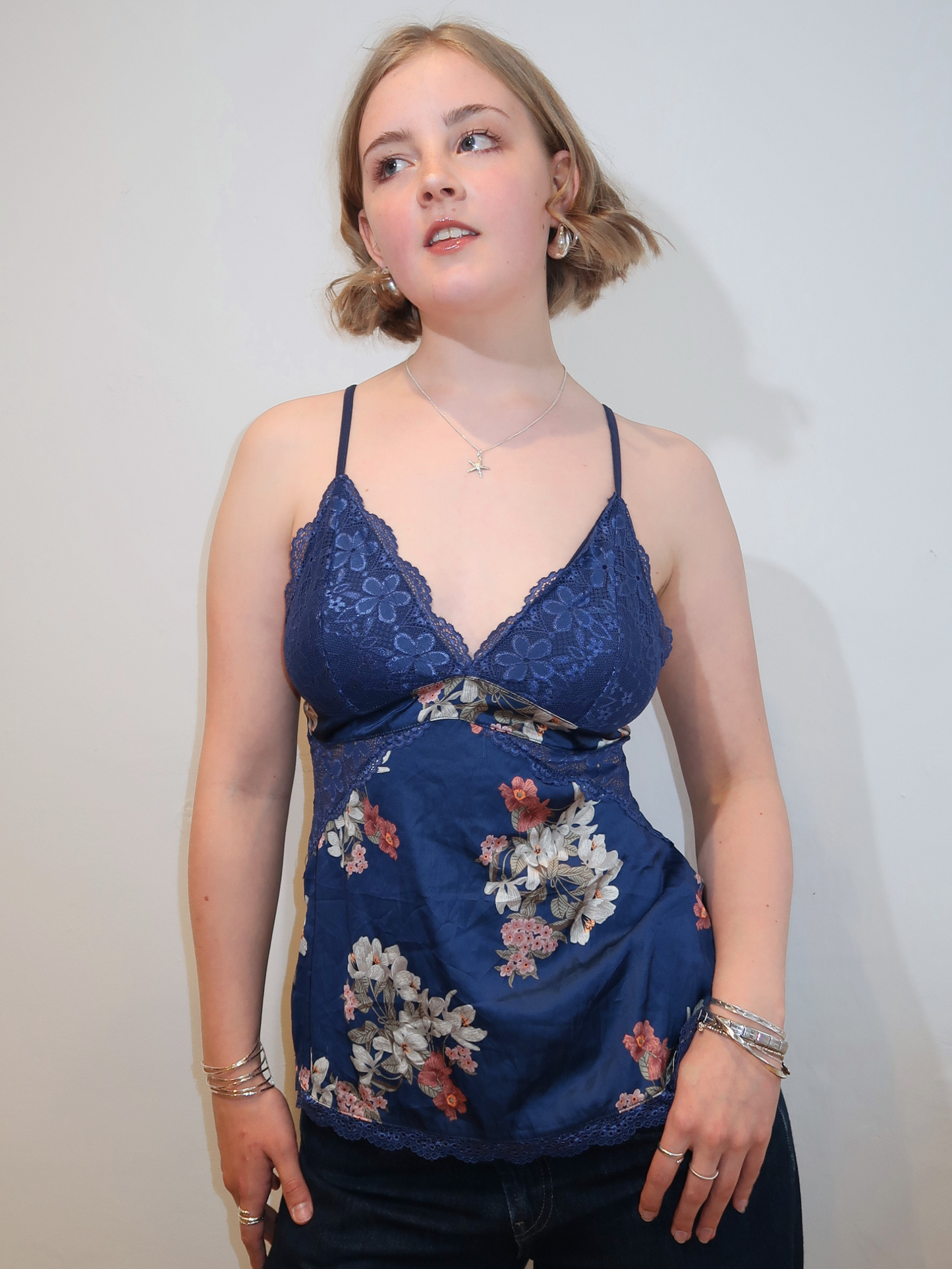 A woman wearing a navy blue lace-trim camisole top with a floral print bodice, paired with dark jeans, standing against a plain off-white wall.