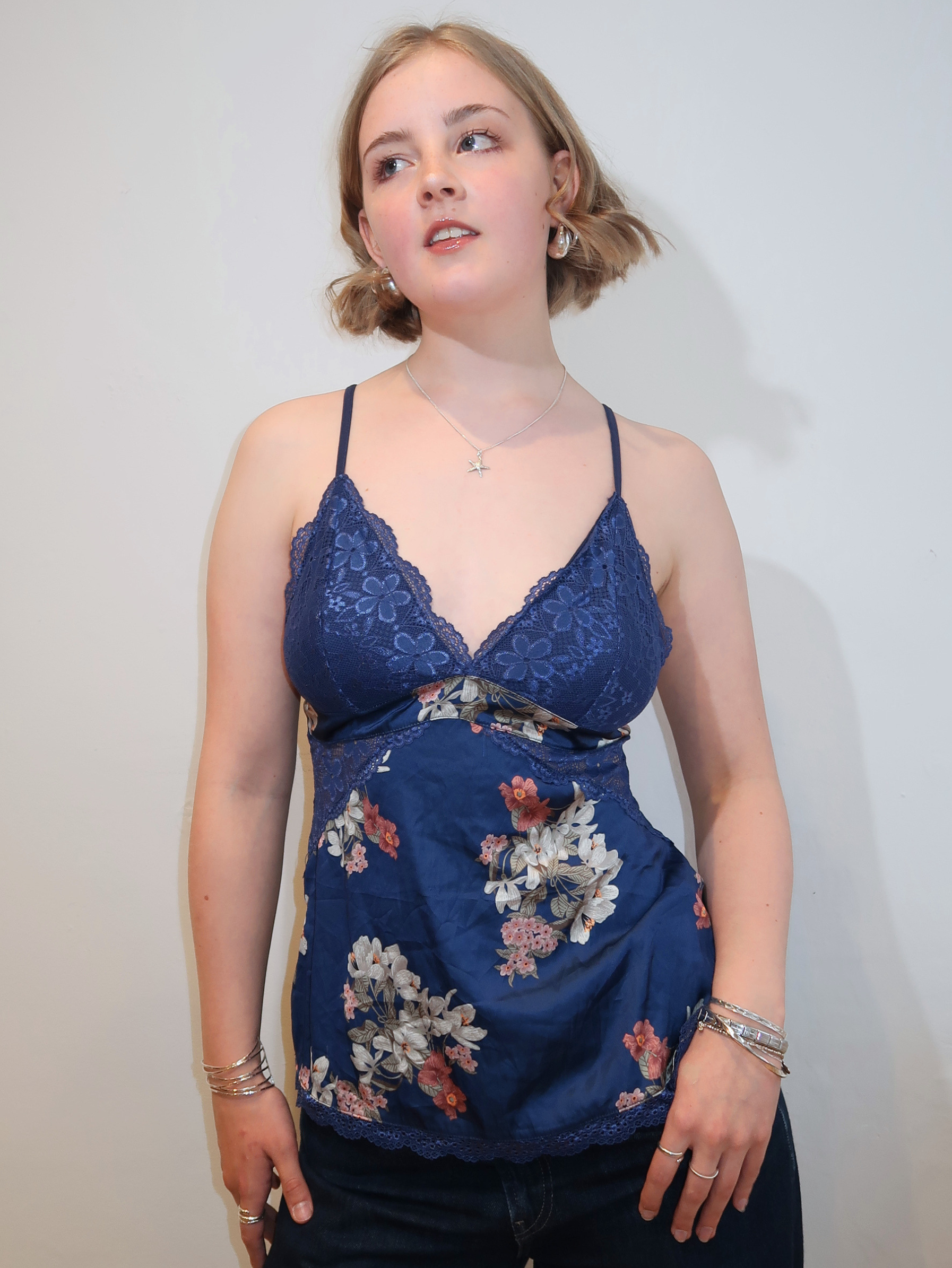 A woman wearing a navy blue lace-trim camisole top with a floral print bodice, paired with dark jeans, standing against a plain off-white wall.