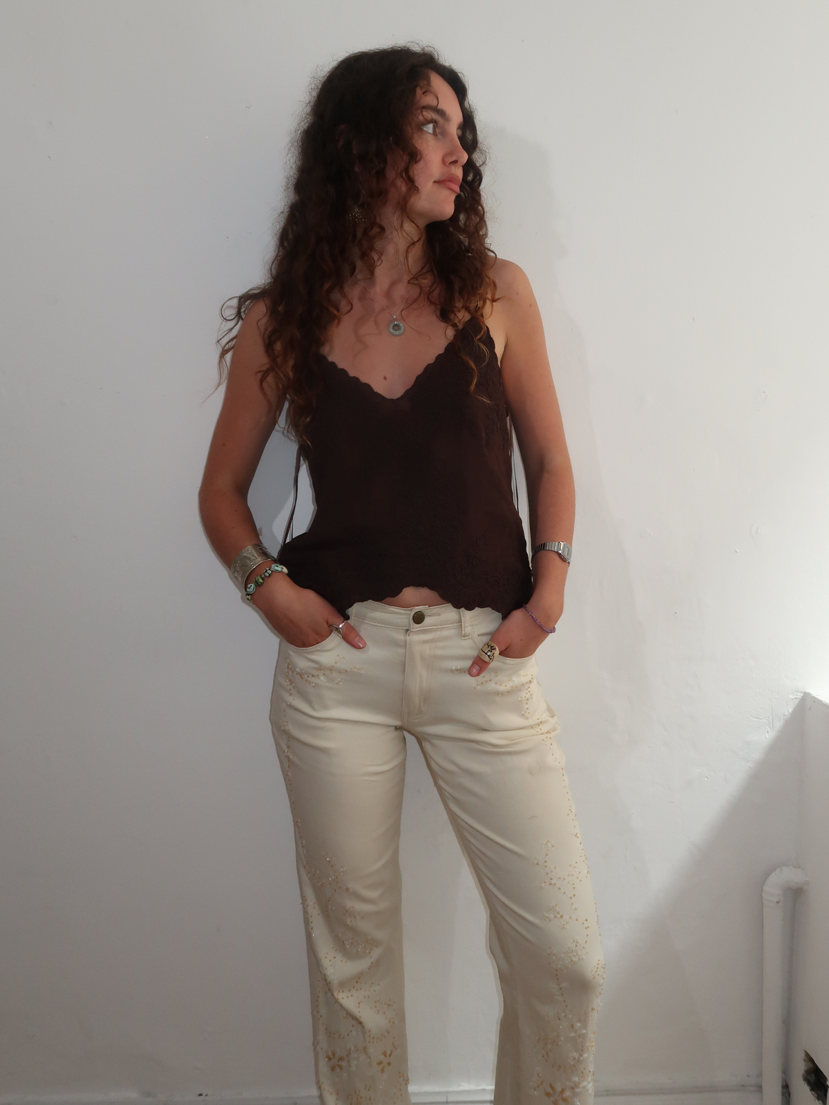 A woman with long curly brown hair stands against a plain white wall wearing a dark brown camisole top and cream-colored jeans with floral embroidery on the lower legs.