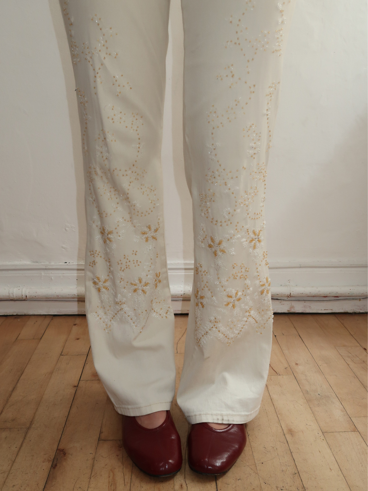 White wide-leg pants with gold floral embroidery down the front of each leg, shown on a model wearing burgundy flats against a white wall and wooden floor.