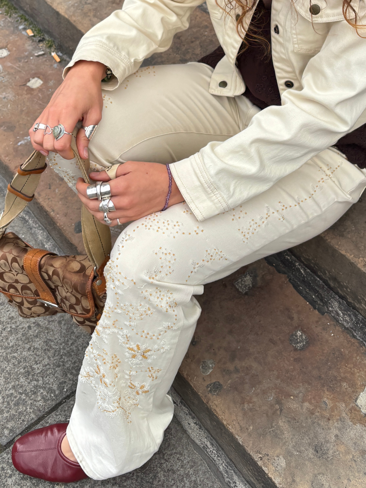A person seated outdoors on stone steps wearing cream-colored embroidered pants, a cream jacket, brown patterned boots, and burgundy shoes, with multiple silver rings on their hands.