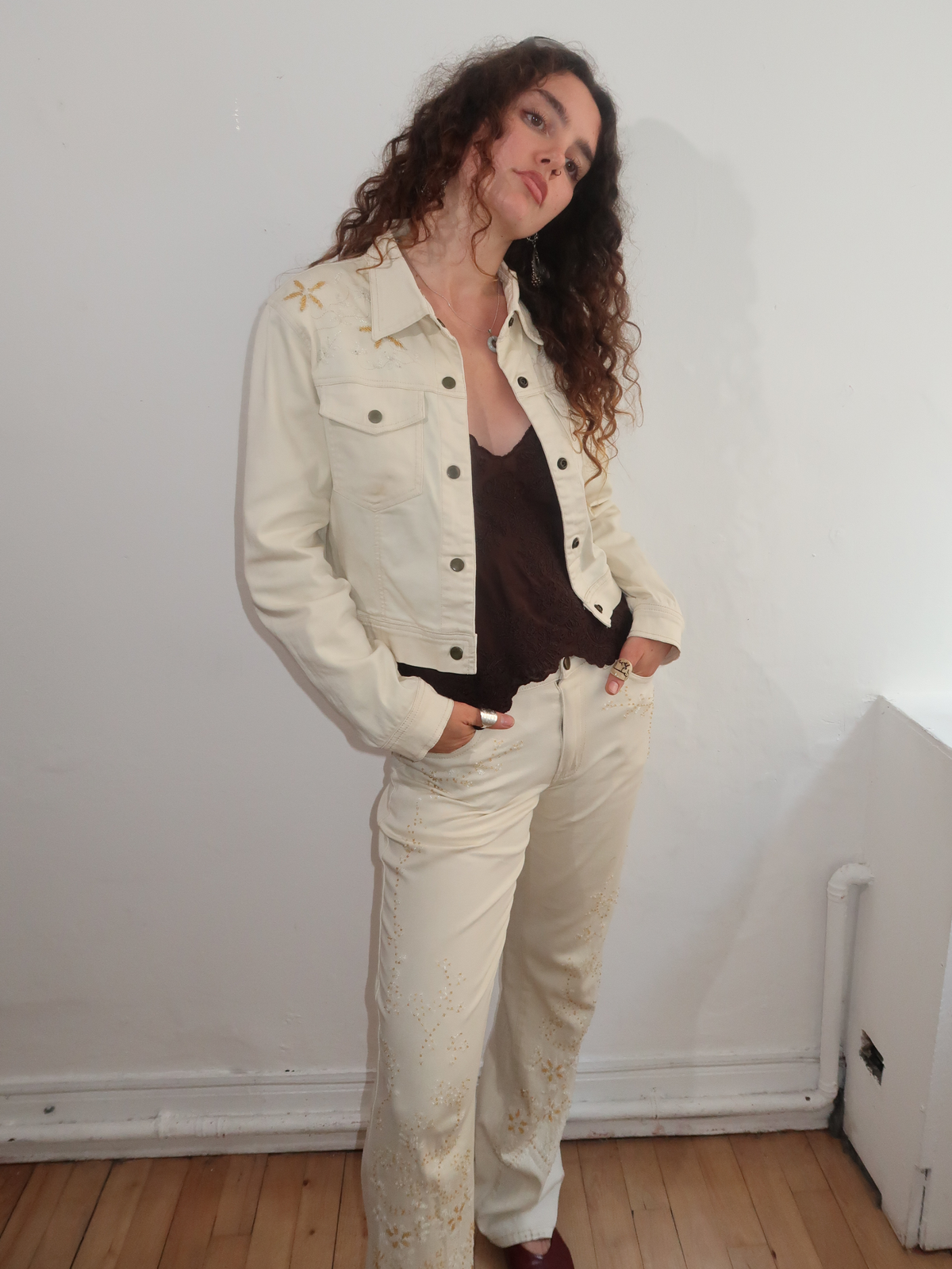 A woman with long curly brown hair stands in a room wearing a cream-colored denim jacket and matching cream pants with gold embroidered details, layered over a dark brown top.