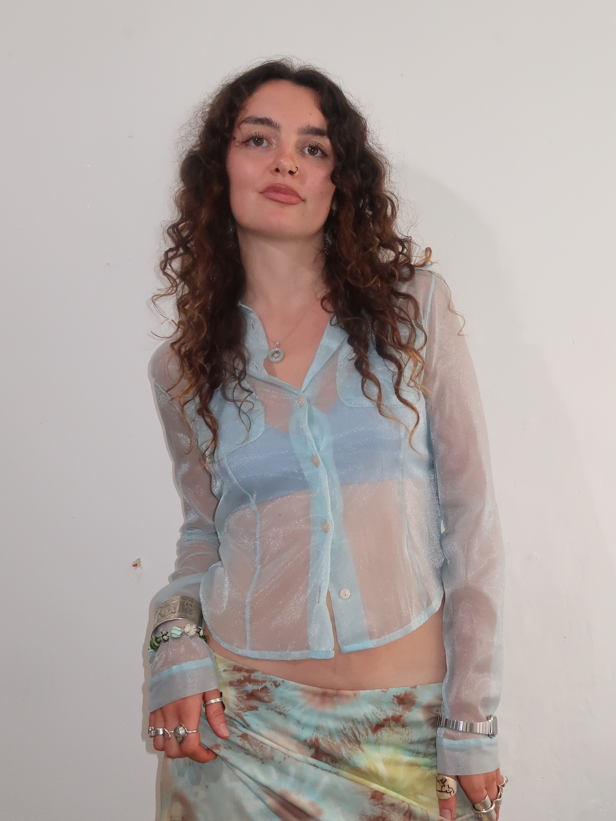 A woman with long, curly brown hair wearing a sheer light blue button-up shirt over a matching light blue top, paired with a tie-dye skirt, standing against a plain off-white wall.