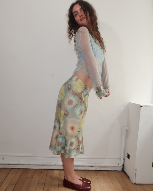 A woman stands in profile wearing a sheer light blue long-sleeve top and a floral midi skirt with a ruffled hem, paired with dark red flats, against a plain white wall and wooden floor.