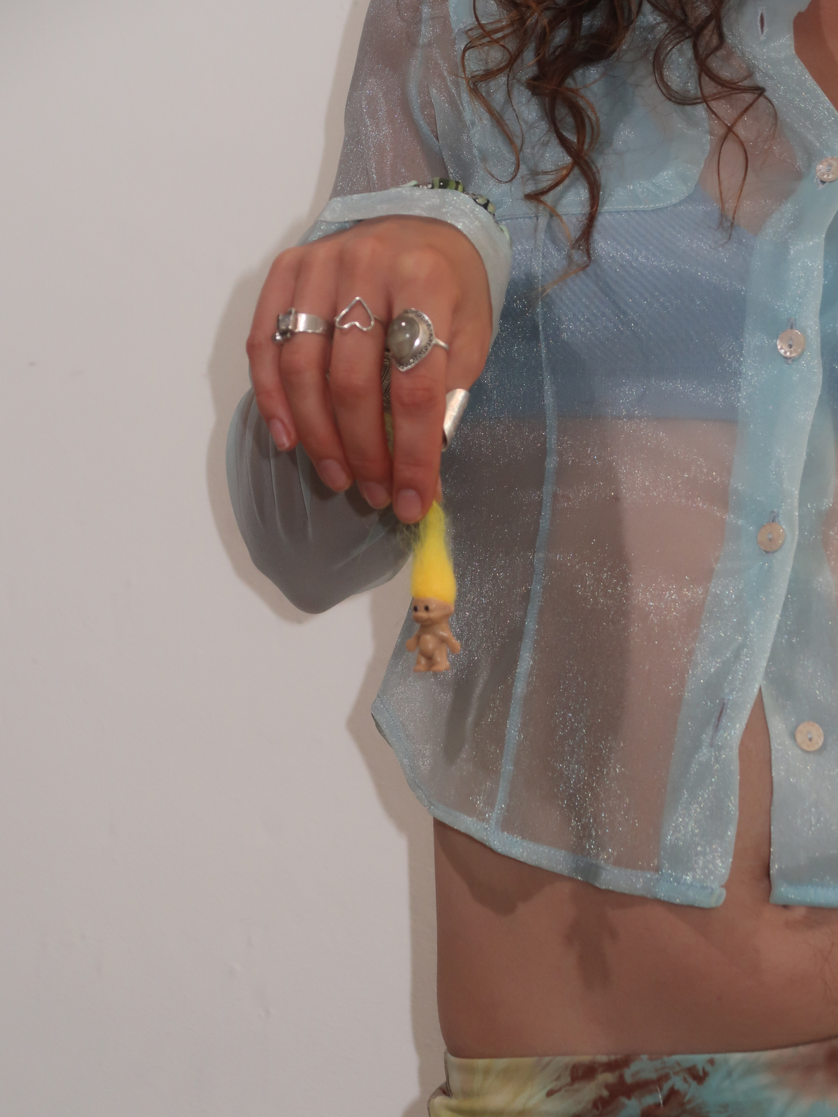 A woman stands in profile wearing a sheer light blue long-sleeve top and a floral midi skirt with a ruffled hem, paired with dark red flats, against a plain white wall and wooden floor.