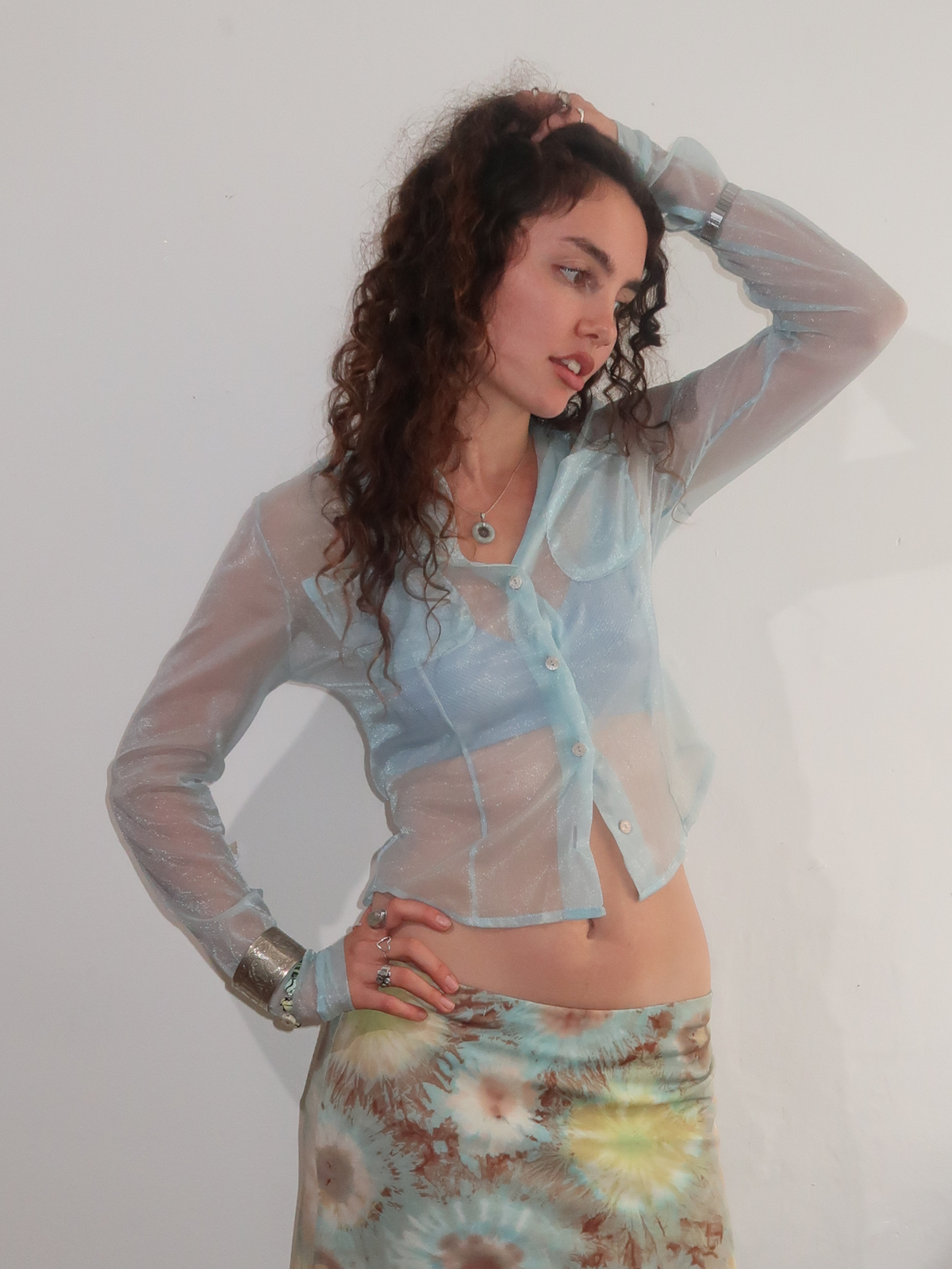 A woman modeling a sheer light blue long-sleeve button-up shirt over a matching light blue bralette, paired with a tie-dye skirt, against a plain white wall.