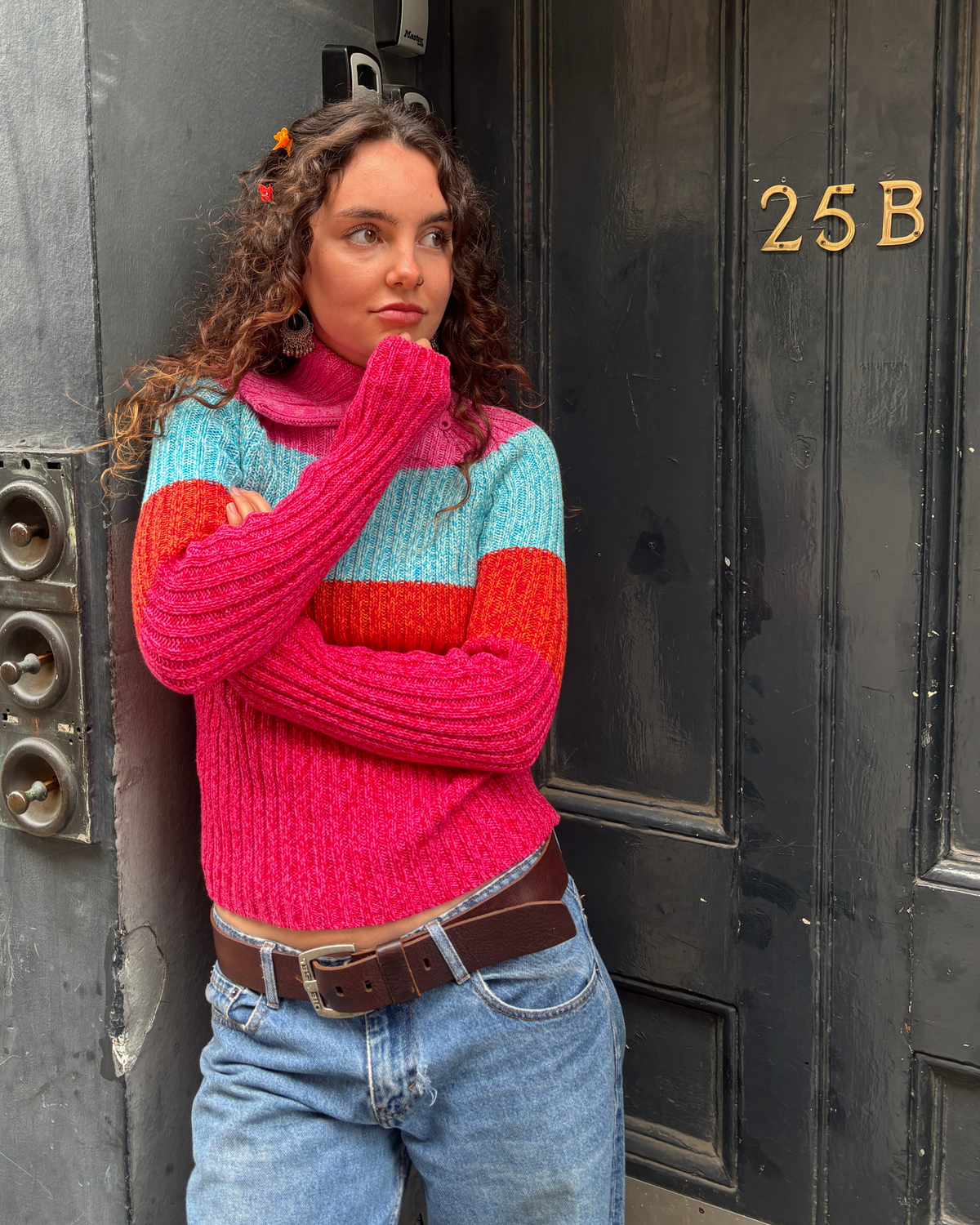 A woman with curly brown hair stands in front of a dark door wearing a chunky knit turtleneck sweater with horizontal stripes in hot pink, light blue, and coral red, paired with light-wash blue jeans and a brown belt.