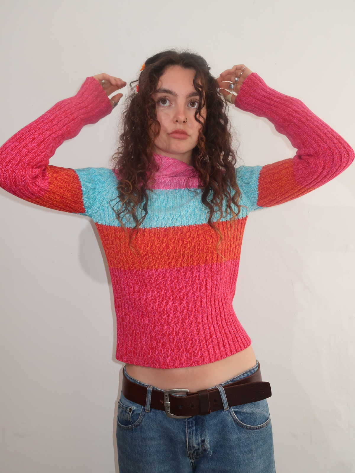 A woman with curly brown hair wearing a colorful striped knit sweater, blue jeans, and a brown belt, posing against a plain white wall.