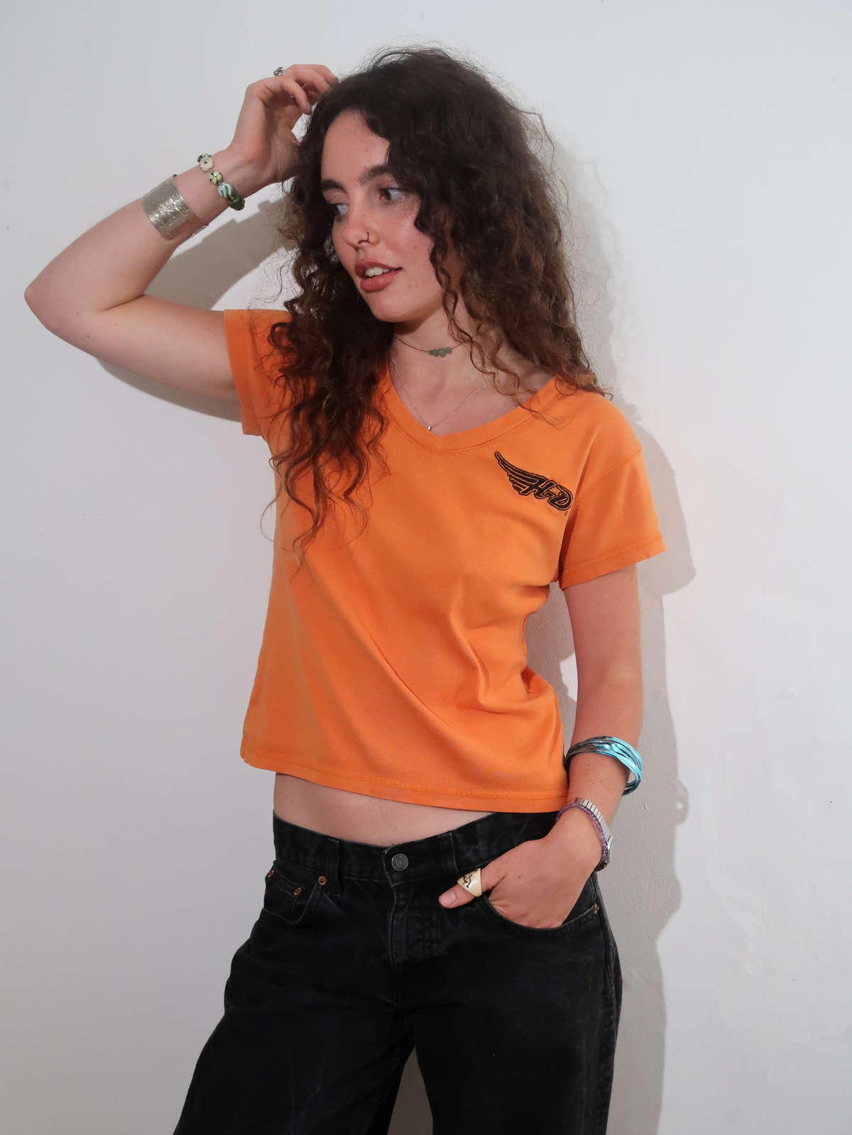 A woman with long curly brown hair wearing a bright orange short-sleeve V-neck t-shirt with a black wing harley davidson graphic on the left chest, paired with black jeans, standing against a plain off-white wall.