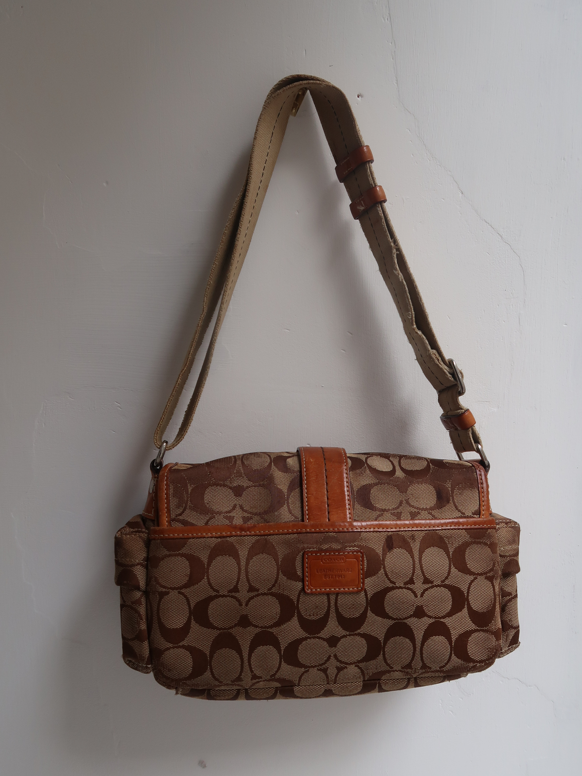 A Coach shoulder bag in the signature monogram canvas, featuring brown and tan tones with a brown leather strap and trim, hanging against a plain light gray wall.